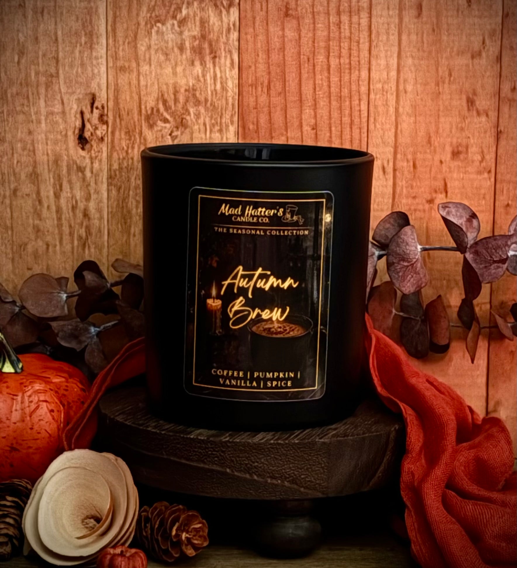 A Fall scented candle in a matte black jar sits on a dark wooden pedestal against a rustic wood background. The candle is surrounded by autumn-themed props including a small orange pumpkin, pinecones, dried leaves, and orange fabric.