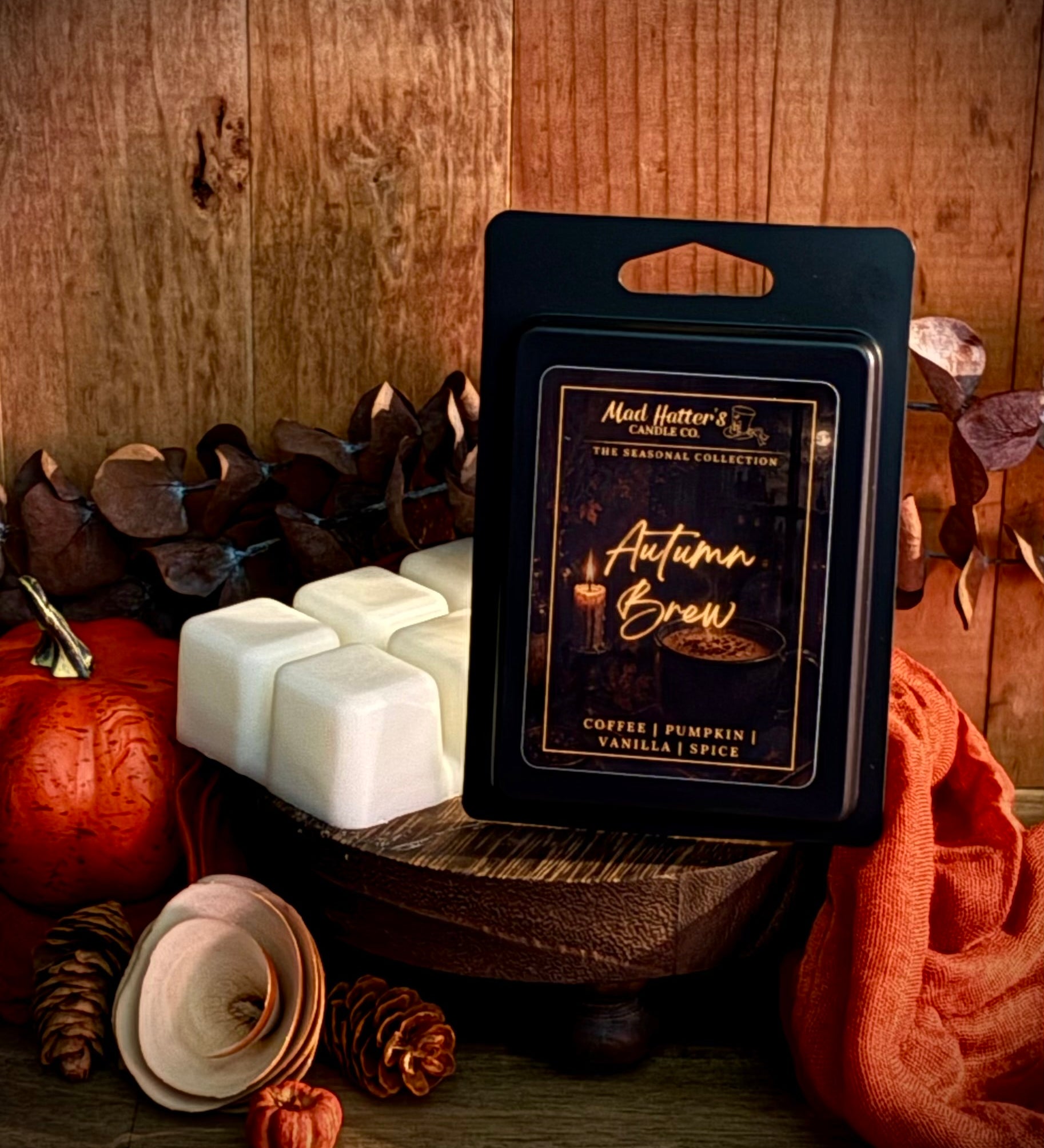 A packaged wax melt product labeled 'Autumn Brew' stands on a wooden surface with several white wax cubes beside it. The scene is styled with pumpkins, pinecones, dried leaves, and an orange fabric against a rustic wood plank background.