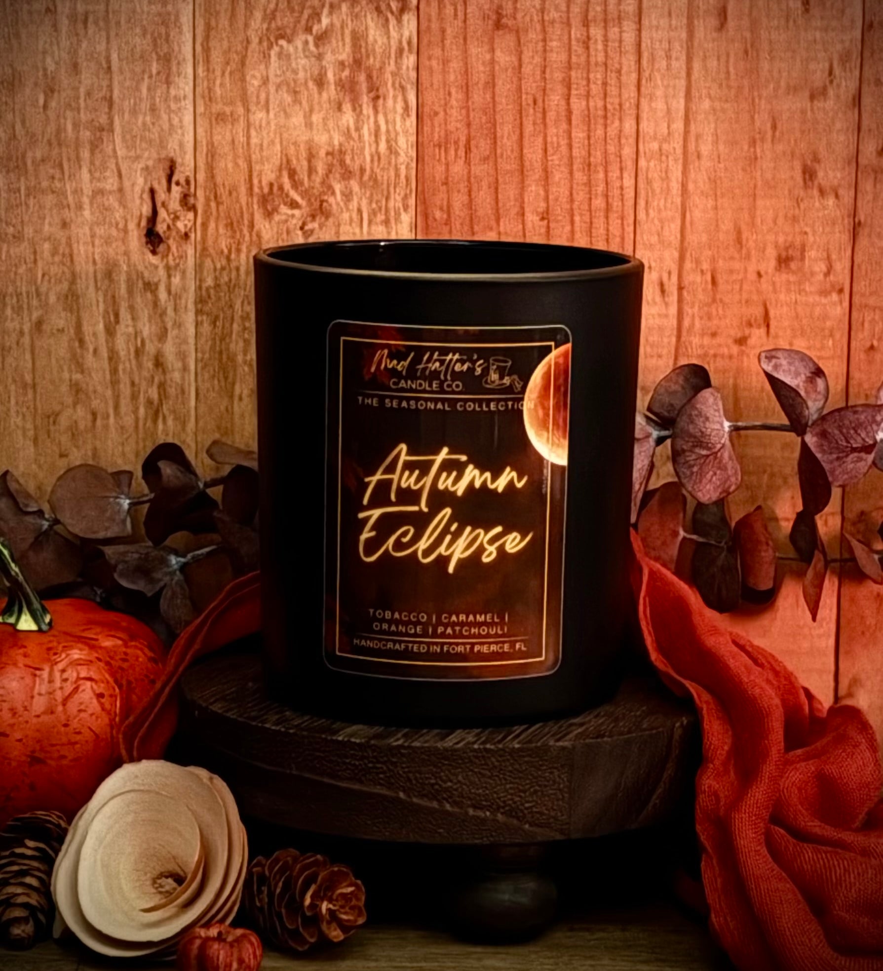 A Fall scented candle in a black jar labeled 'Autumn Eclipse' is styled on a wooden pedestal with autumn props including a pumpkin, dried leaves, pinecones, and orange fabric against a rustic wood background.