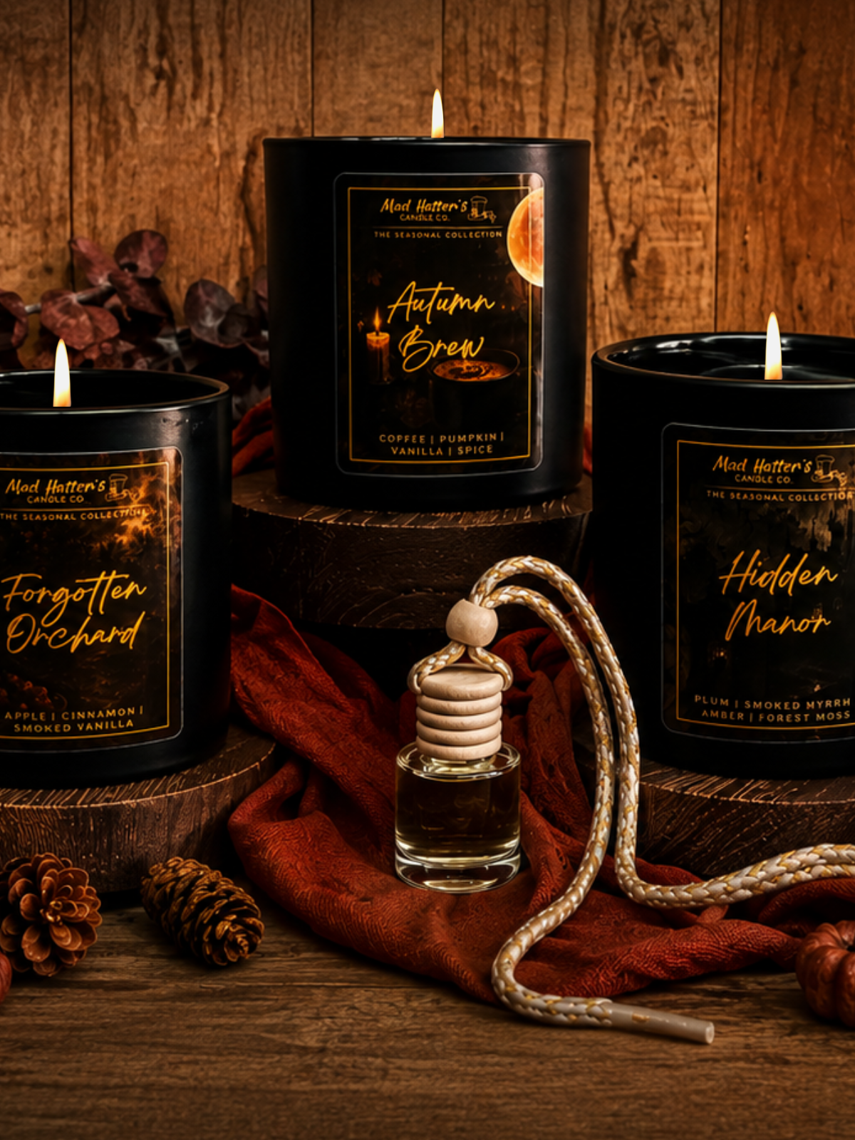 Four scented Fall candles in black jars, along with wax melts, and a car freshener are arranged on a rustic wooden surface with autumn-themed props including a small pumpkin, pinecones, dried leaves, and orange fabric. The candle labels read Autumn Brew, Forgotten Orchard, Hidden Manor, and Autumn Eclipse.