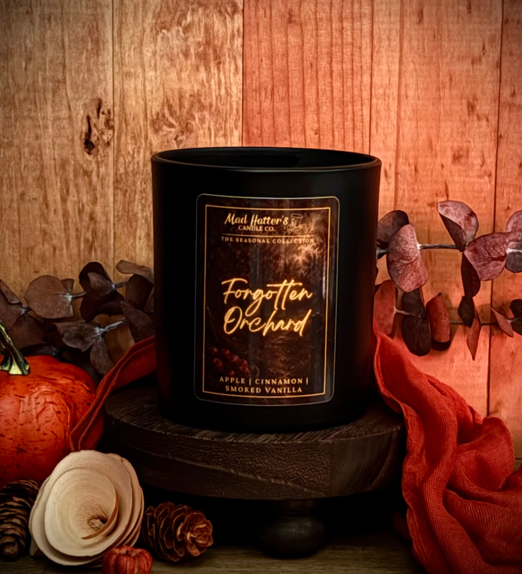 A Fall candle in a black jar with a label reading 'Forgotten Orchard' sits on a dark wooden pedestal against a rustic wood background, surrounded by autumn props including a pumpkin, dried leaves, pinecones, and orange fabric.