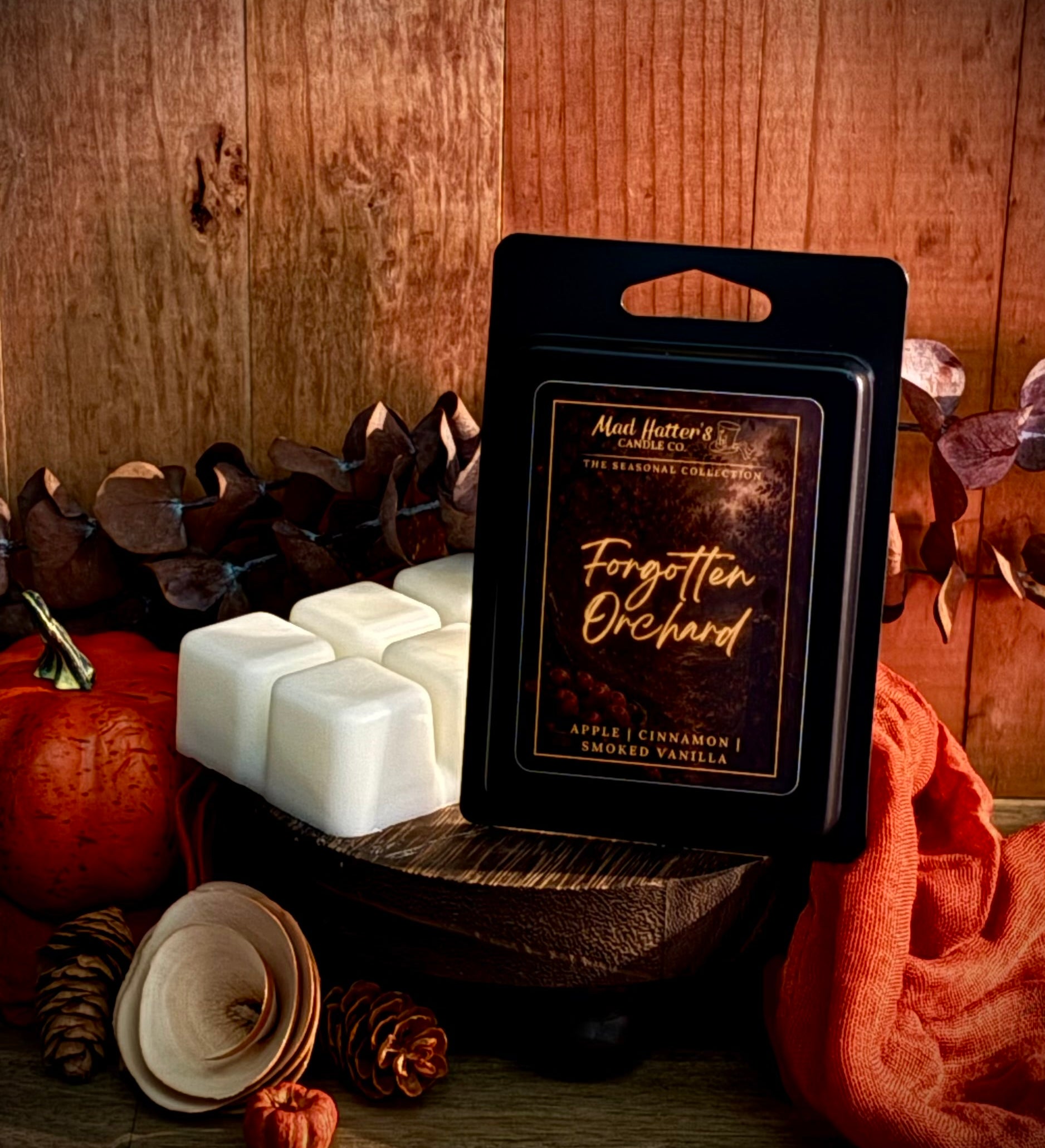 A packaged wax melt labeled 'Forgotten Orchard' sits on a wooden surface with several white wax cubes beside it. The scene is styled with autumn props including a pumpkin, pinecones, dried leaves, and orange fabric against a wood plank background.