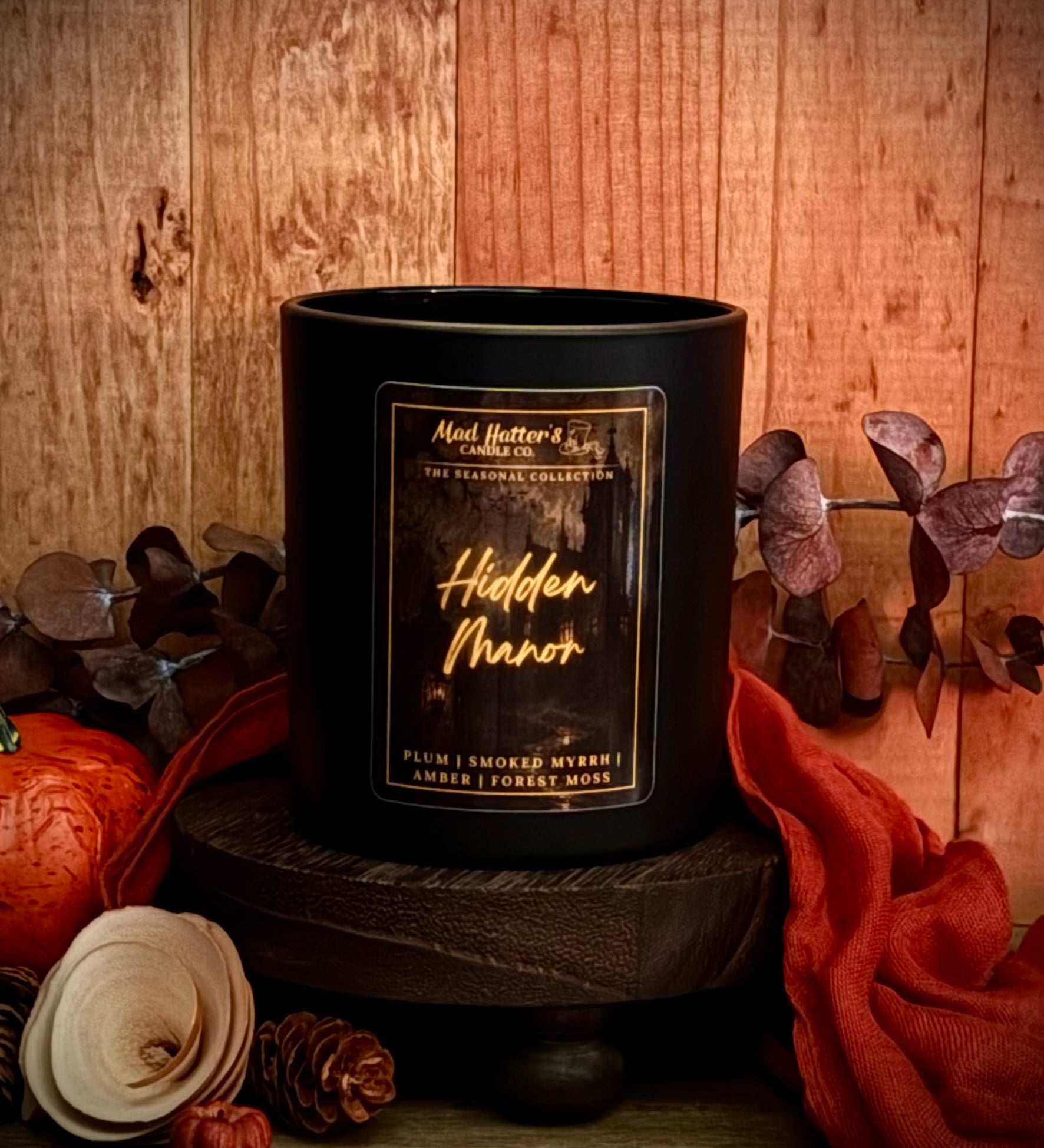 A Fall scented candle in a black jar with label reading 'Hidden Manor' sits on a dark wooden pedestal against a rustic wood background, styled with autumn props including a pumpkin, dried leaves, pinecones, and orange fabric.