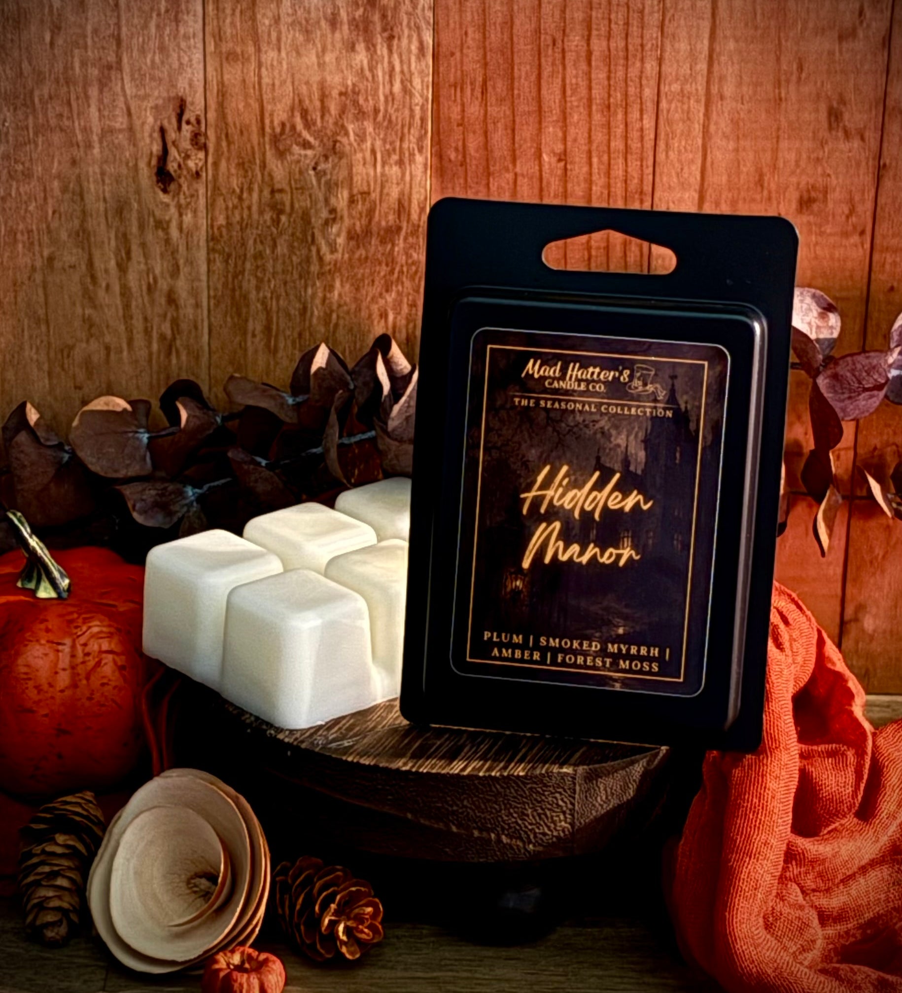 A package of wax melts stands on a wooden surface beside several white wax cubes, with autumn props including a pumpkin, pinecones, dried leaves, and an orange cloth against a rustic wood plank background. The package label reads 'Hidden Manor' 
