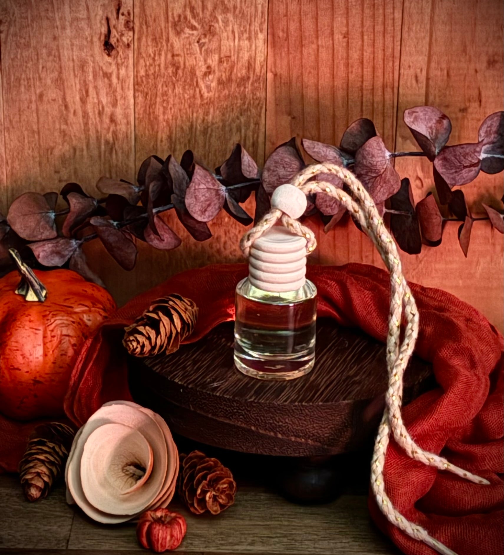 A small glass bottle car freshener sits on a dark wooden pedestal, surrounded by autumn props including an orange cloth, pinecones, dried leaves, and a pumpkin against a wood plank background.